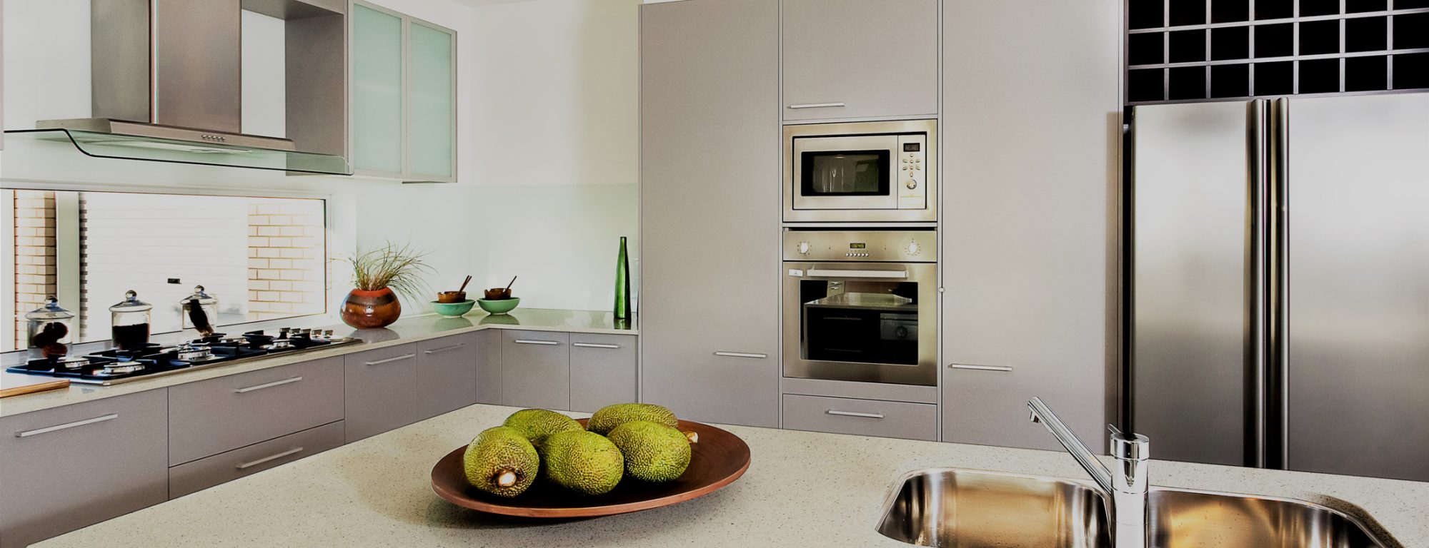 Counter top made in ceramic seen closely with fruits on a brown color clay dish next to the modern  silver faucet attached to the sink, the silver refrigerator and the wall oven with pantry cupboards next to the gas stove